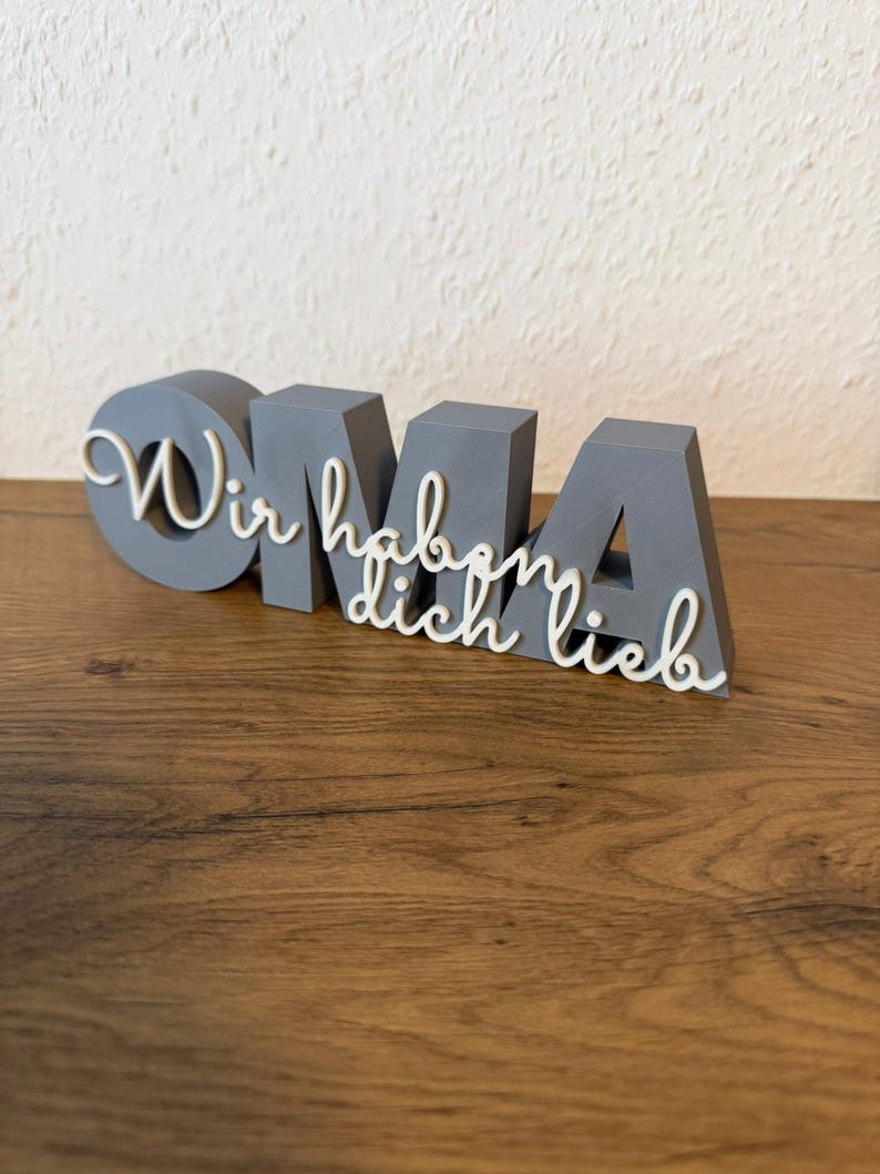 K&ouml;nnte beinhalten: Ein graues, 3D-gedrucktes Schild mit dem Wort "OMA" in Blockbuchstaben, mit dem kursiven Text "Wir haben dich lieb" dar&uuml;ber. Das Schild steht auf einer Holzoberfl&auml;che.