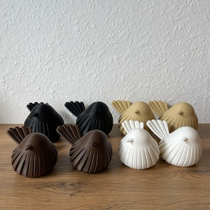 Eine Nahaufnahme zeigt acht moderne, gerippte Vogelskulpturen im minimalistischen Japandi-Stil, gefertigt aus umweltfreundlichem 3D-Druck-Material. Sie sind in vier Paaren auf einer Holzoberfläche angeordnet: ein Paar in Matt-Dunkelgrau, ein Paar in Sand-Beige, ein Paar in Schokobraun und ein Paar in Reinweiß.