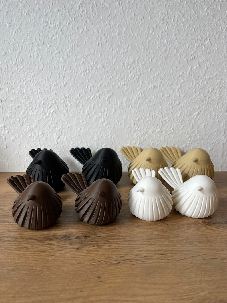 Eine Nahaufnahme zeigt acht moderne, gerippte Vogelskulpturen im minimalistischen Japandi-Stil, gefertigt aus umweltfreundlichem 3D-Druck-Material. Sie sind in vier Paaren auf einer Holzoberfläche angeordnet: ein Paar in Matt-Dunkelgrau, ein Paar in Sand-Beige, ein Paar in Schokobraun und ein Paar in Reinweiß.