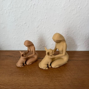 May include: Two beige monochromatic figurines, each depicting a kneeling person with long hair gently holding a small dog. The figures are smooth, featureless, and minimalist in form, showcasing a soft matte finish and a consistent light tan color.