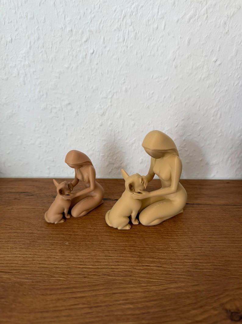 May include: Two beige monochromatic figurines, each depicting a kneeling person with long hair gently holding a small dog. The figures are smooth, featureless, and minimalist in form, showcasing a soft matte finish and a consistent light tan color.
