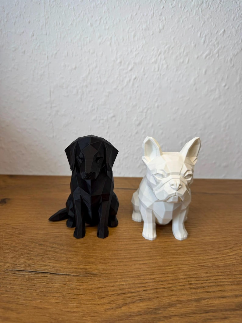 May include: Two geometric dog figurines, one black and one white, featuring a faceted, low-poly design. The black figurine resembles a Labrador Retriever, while the white figurine resembles a French Bulldog, both sitting in an upright, alert posture.