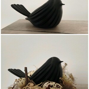 May include: A matte black bird figurine with a ribbed texture on its body and a pointed beak. The second view shows the same black bird figurine nestled inside a small bird's nest filled with speckled cream-colored eggs and brown feathers.