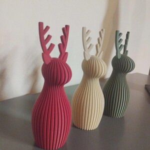 May include: Three standing deer figurines with a vertical ribbed texture. The first is deep red, the second is cream, and the third is sage green. Each deer has a rounded body and prominent antlers extending from the top of the head.