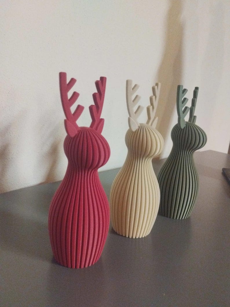 May include: Three standing deer figurines with a vertical ribbed texture. The first is deep red, the second is cream, and the third is sage green. Each deer has a rounded body and prominent antlers extending from the top of the head.
