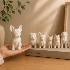 May include: A collection of seven white, geometric-style dog figurines in various breeds, including a French Bulldog, English Bulldog, and others, displayed on a book titled KINFOLK, with one figurine held in a hand.