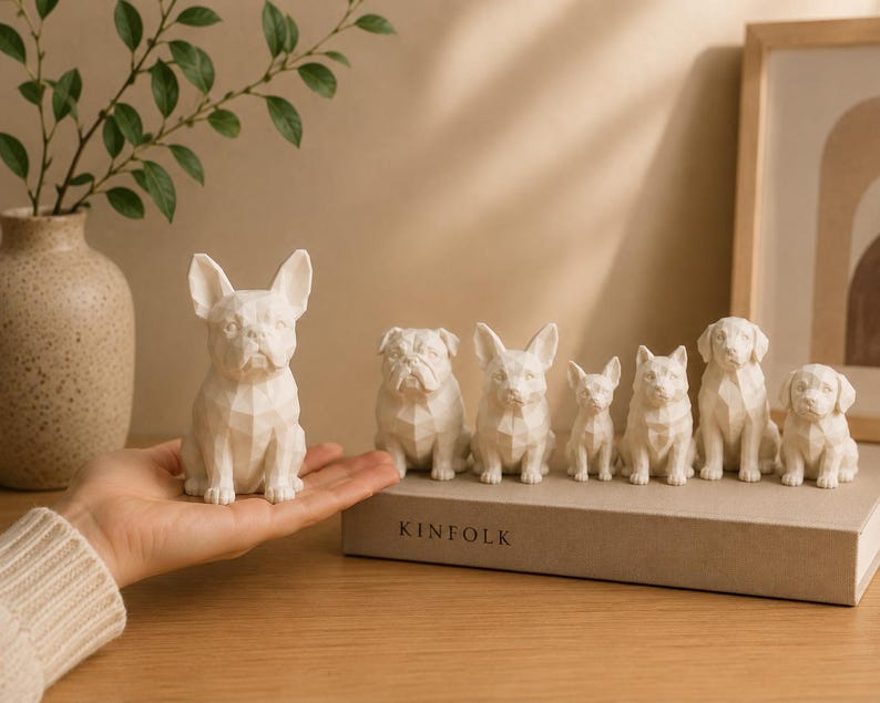 May include: A collection of seven white, geometric-style dog figurines in various breeds, including a French Bulldog, English Bulldog, and others, displayed on a book titled KINFOLK, with one figurine held in a hand.