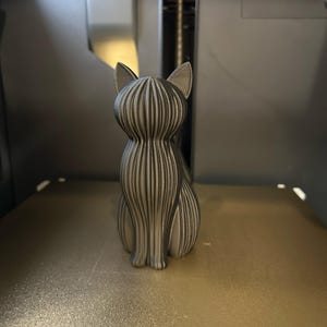 May include: A 3D printed cat figurine in a metallic silver finish, featuring a distinct vertical ribbed texture across its entire body, pointed ears, and a seated posture, captured from a rear-facing perspective.