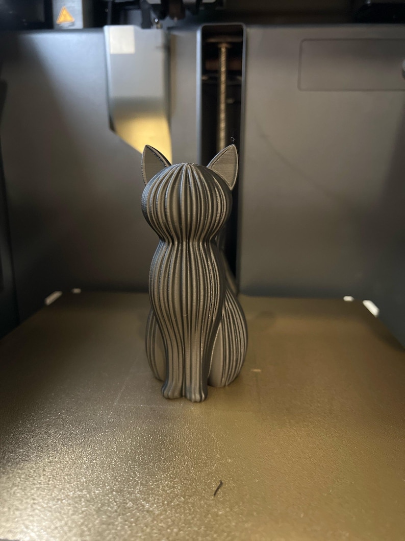 May include: A 3D printed cat figurine in a metallic silver finish, featuring a distinct vertical ribbed texture across its entire body, pointed ears, and a seated posture, captured from a rear-facing perspective.