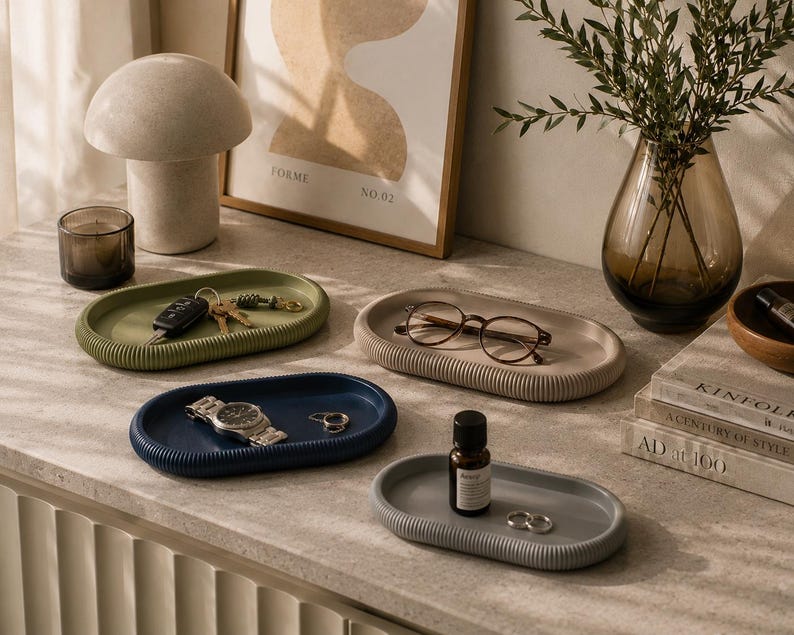 Puede incluir: Una colecci&oacute;n de bandejas ovaladas decorativas en varios colores, incluyendo verde, azul y gris. Cada bandeja tiene una textura acanalada y contiene objetos como llaves, gafas, un reloj y peque&ntilde;as botellas. Tambi&eacute;n se ve un adorno en forma de hongo y un jarr&oacute;n.
