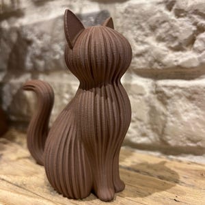 May include: A brown cat figurine featuring a distinct ribbed or layered texture across its entire body. The cat is depicted in a seated position with its tail curved upward, showcasing a vertical linear pattern that defines its form.