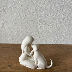 May include: A matte white figurine depicting a stylized human figure kneeling and interacting with a dog. The human figure has long hair and is positioned facing the dog, with one hand gently touching the dog's head. The dog is sitting upright.