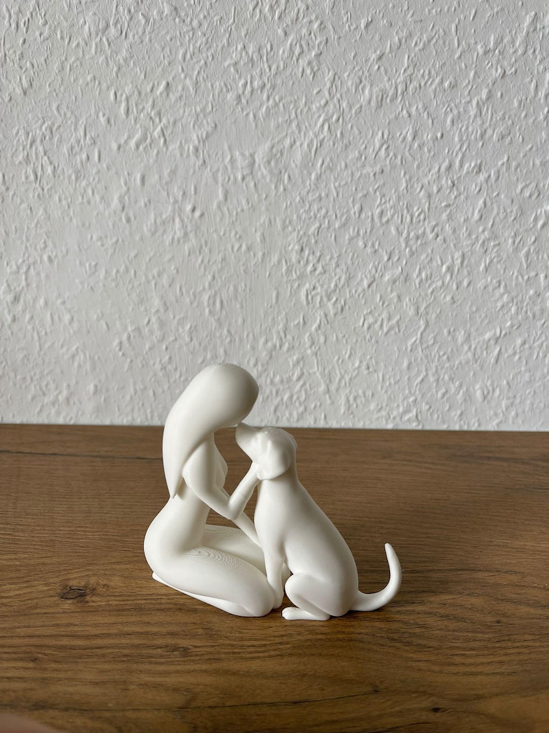 May include: A matte white figurine depicting a stylized human figure kneeling and interacting with a dog. The human figure has long hair and is positioned facing the dog, with one hand gently touching the dog's head. The dog is sitting upright.