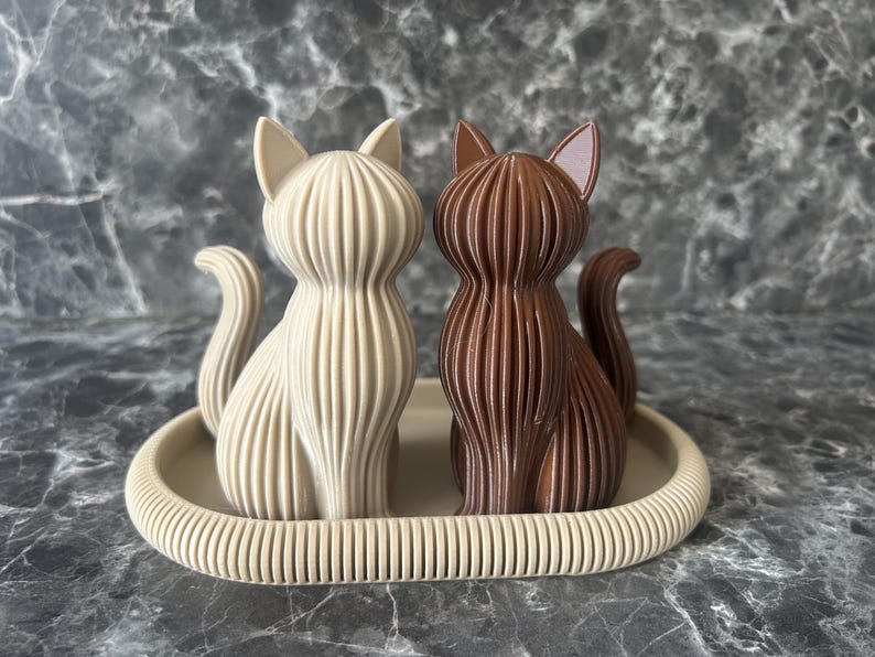 Zwei moderne Katzen-Skulpturen aus nachhaltigem PLA im minimalistischen Design. Das Duo in Wüstenbraun und Dunkel-Schokobraun zeigt die feine gerippte Textur des 3D-Drucks und ist auf einer farblich abgestimmten Schale arrangiert. Ideal als skandinavische Tischdeko.
Die Schale ist nicht im Bestellumfang enthalten.