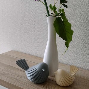 May include: A tall white ceramic vase containing a purple allium flower and green leaves, positioned next to two small bird figurines. One bird is matte gray and the other is cream-colored, both featuring a ribbed, layered wing design.