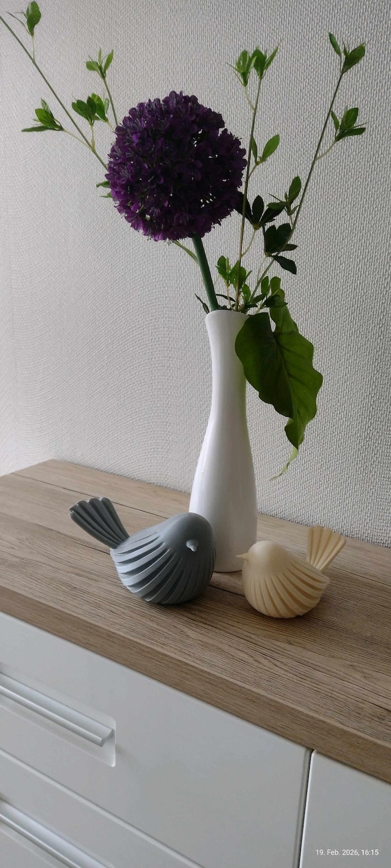 May include: A tall white ceramic vase containing a purple allium flower and green leaves, positioned next to two small bird figurines. One bird is matte gray and the other is cream-colored, both featuring a ribbed, layered wing design.