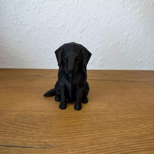May include: A black dog figurine crafted with a geometric, low-poly design. The sculpture features sharp, angular facets forming the shape of a sitting canine with floppy ears, positioned in a forward-facing orientation on a flat surface.