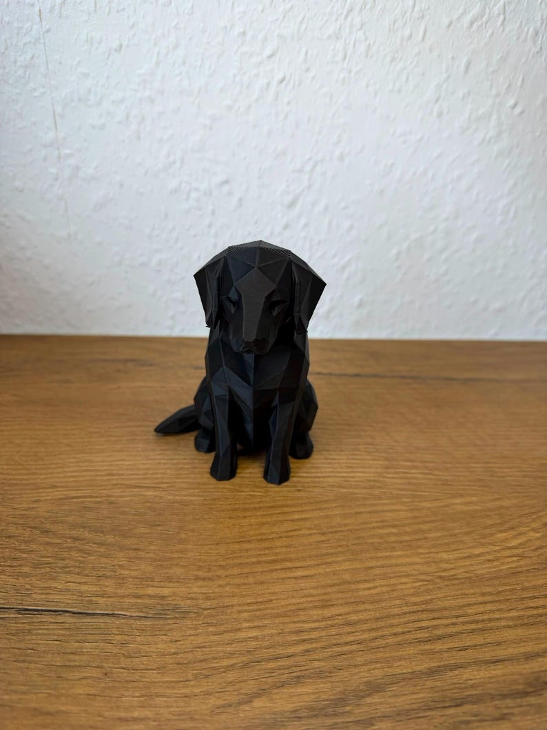 May include: A black dog figurine crafted with a geometric, low-poly design. The sculpture features sharp, angular facets forming the shape of a sitting canine with floppy ears, positioned in a forward-facing orientation on a flat surface.