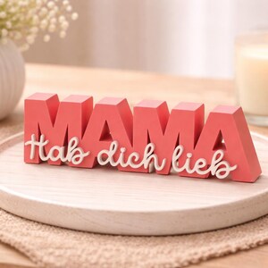 K&ouml;nnte beinhalten: Ein korallenfarbenes Holzschild mit dem Schriftzug "MAMA" in Blockbuchstaben und den Worten "Hab dich lieb" in Schreibschrift darunter. Das Schild steht auf einem Holztablett, im Hintergrund eine Kerze und Blumen.
