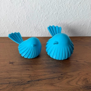 May include: Two bright blue bird figurines with rounded bodies and fanned tail feathers. Each bird features a smooth, matte finish and a small, pointed beak. The figures are positioned side-by-side, displaying a consistent vibrant color and texture.