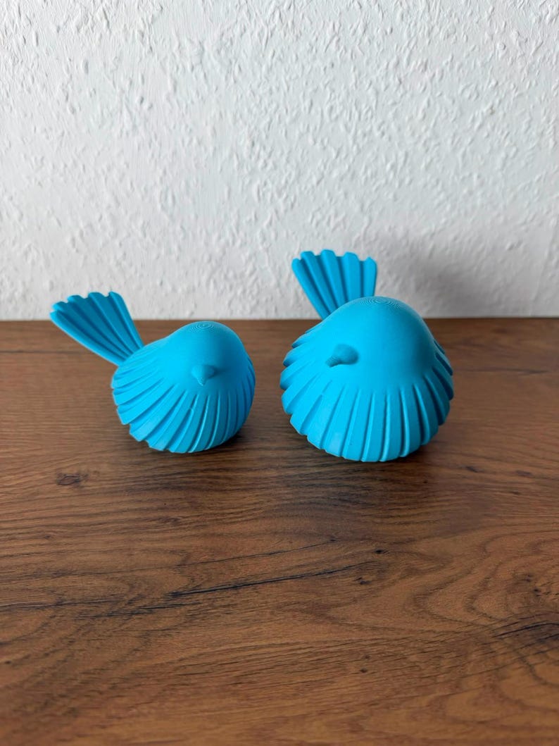 May include: Two bright blue bird figurines with rounded bodies and fanned tail feathers. Each bird features a smooth, matte finish and a small, pointed beak. The figures are positioned side-by-side, displaying a consistent vibrant color and texture.