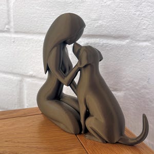 May include: A matte dark gray sculpture depicting a stylized figure kneeling and gently touching the head of a dog. The smooth, minimalist form captures a moment of connection between the human-like silhouette and the canine companion.