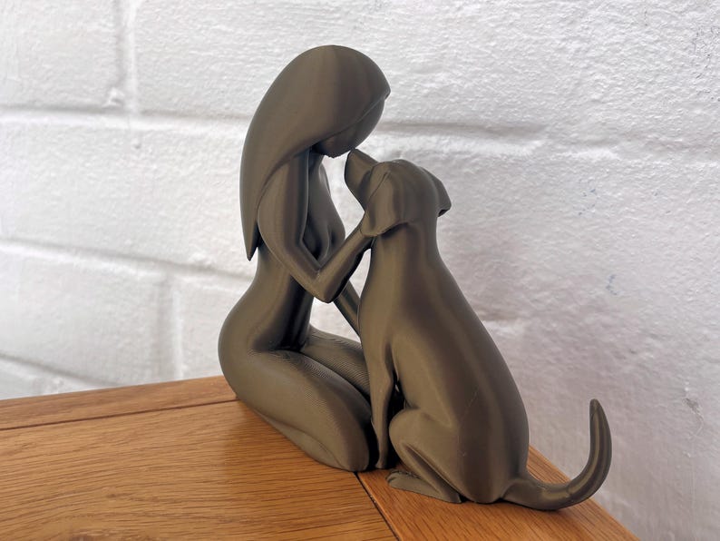May include: A matte dark gray sculpture depicting a stylized figure kneeling and gently touching the head of a dog. The smooth, minimalist form captures a moment of connection between the human-like silhouette and the canine companion.