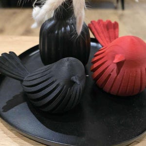 May include: A black circular tray holding two bird-shaped figurines and a small vase. One bird figurine is matte black and the other is matte red. Both birds feature a ribbed, layered texture. A black vase contains white dried decorative grass.