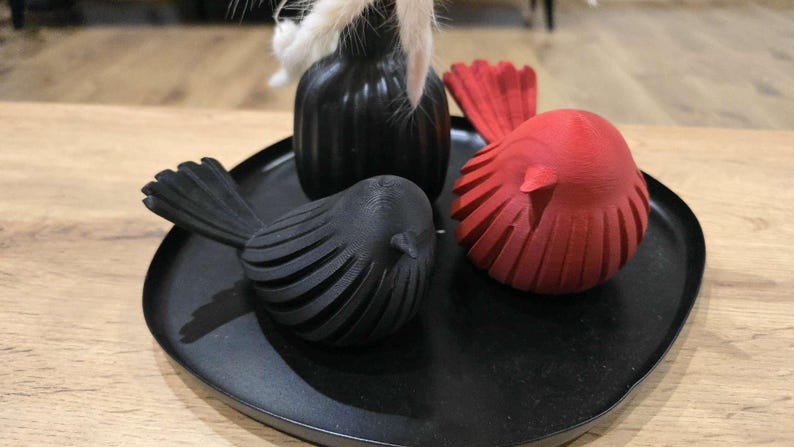 May include: A black circular tray holding two bird-shaped figurines and a small vase. One bird figurine is matte black and the other is matte red. Both birds feature a ribbed, layered texture. A black vase contains white dried decorative grass.
