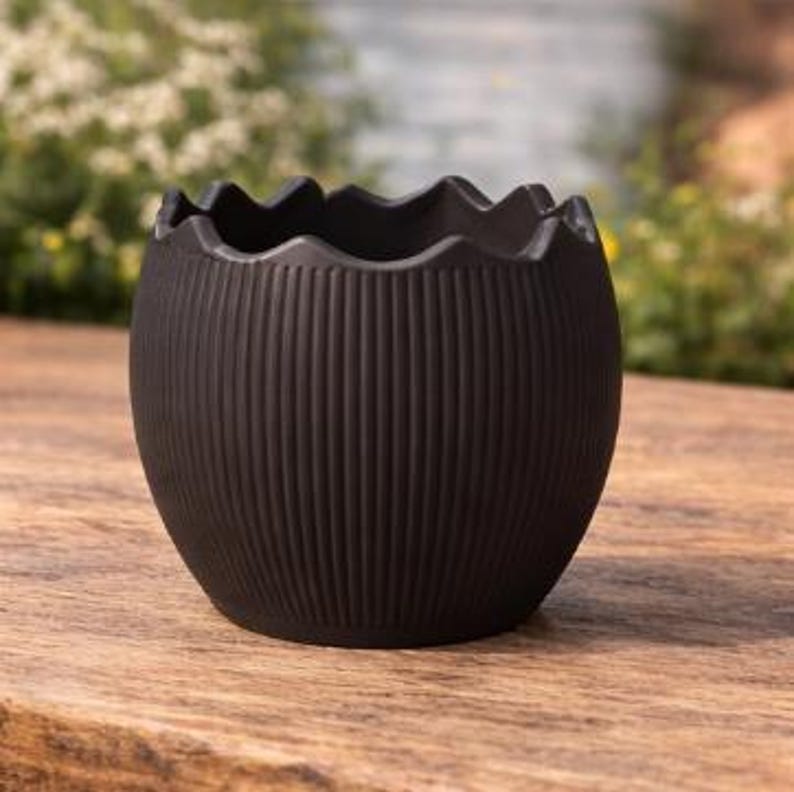 May include: A dark brown, egg-shaped ceramic planter with a textured, ribbed exterior and a wavy rim. The planter sits on a wooden surface, with a blurred background of greenery.