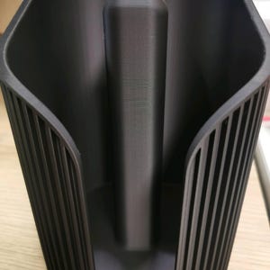 May include: A matte black plastic paper towel holder featuring a central vertical rod for holding a roll and a curved, ribbed exterior shell design that provides structural support while keeping the paper towel roll contained.