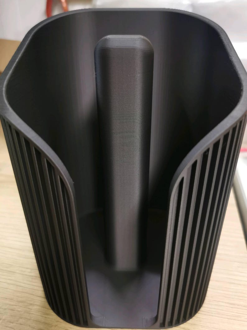 May include: A matte black plastic paper towel holder featuring a central vertical rod for holding a roll and a curved, ribbed exterior shell design that provides structural support while keeping the paper towel roll contained.