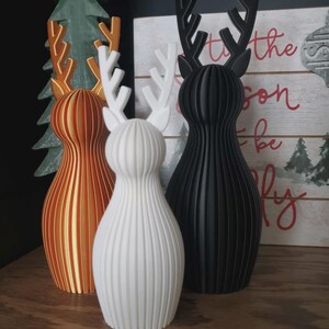 May include: Three 3D-printed reindeer figurines with vertical ribbed textures. One is metallic gold, one is matte white, and one is matte black. Each features stylized antlers and a rounded, tapered body shape standing upright on a flat surface.