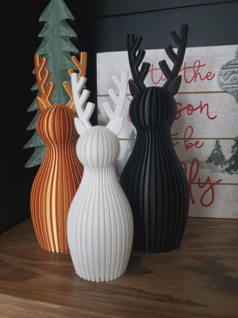 May include: Three 3D-printed reindeer figurines with vertical ribbed textures. One is metallic gold, one is matte white, and one is matte black. Each features stylized antlers and a rounded, tapered body shape standing upright on a flat surface.