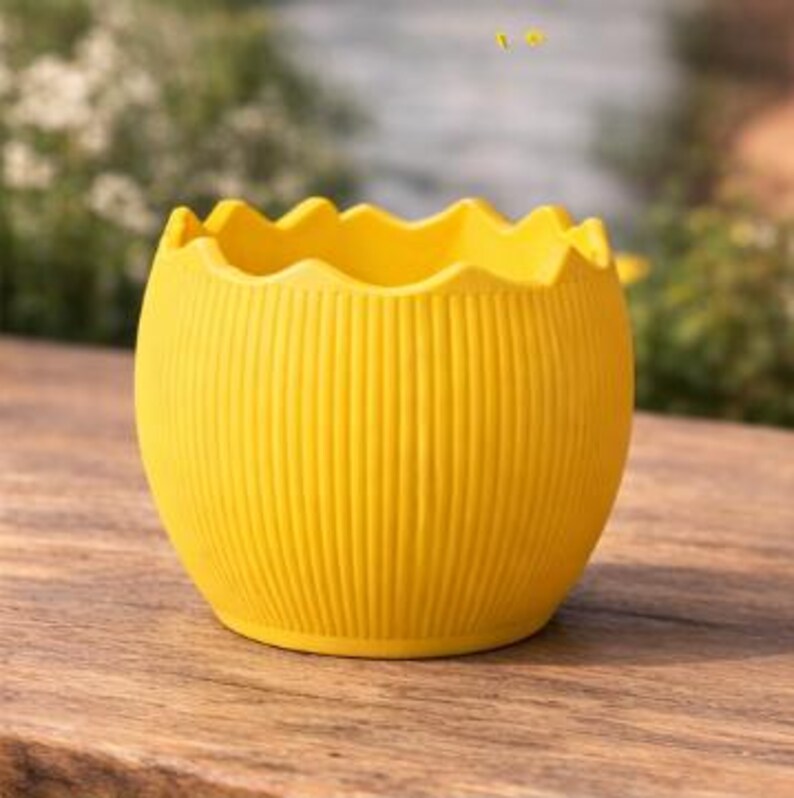 May include: A vibrant yellow, egg-shaped planter with a scalloped rim and vertical ribbed detailing. The planter is positioned on a wooden surface, with a blurred green backdrop implying an outdoor location. The planter is approximately 13 cm in diameter.