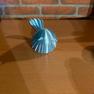 May include: A small, metallic light blue bird figurine with a rounded body and a fanned tail. The surface of the bird features vertical ridges that extend from the center of the back toward the edges, creating a textured, symmetrical appearance.
