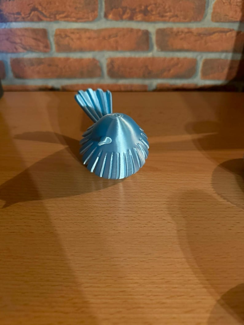May include: A small, metallic light blue bird figurine with a rounded body and a fanned tail. The surface of the bird features vertical ridges that extend from the center of the back toward the edges, creating a textured, symmetrical appearance.