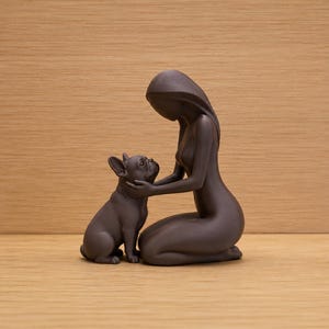 May include: A dark brown matte figurine depicting a kneeling person with long hair gently cupping the face of a French Bulldog. The sculpture features smooth, minimalist curves and a solid, monochromatic finish, capturing a quiet interaction.