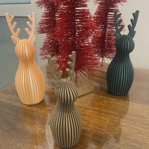 May include: Three 3D-printed reindeer figurines with vertical ribbed textures, featuring one light peach, one metallic bronze, and one dark green deer, positioned in front of two red bottle brush trees on a wooden surface.