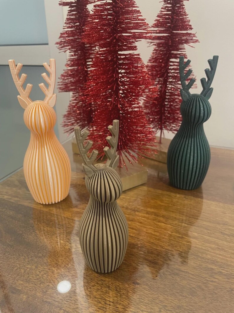 May include: Three 3D-printed reindeer figurines with vertical ribbed textures, featuring one light peach, one metallic bronze, and one dark green deer, positioned in front of two red bottle brush trees on a wooden surface.