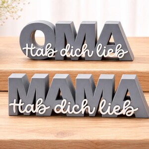 K&ouml;nnte beinhalten: Graue Holzbuchstabenbl&ouml;cke mit den W&ouml;rtern "OMA" und "MAMA" und dem wei&szlig;en Schriftzug "Hab dich lieb". Die Bl&ouml;cke stehen auf einer Holzoberfl&auml;che, im Hintergrund eine Vase mit wei&szlig;en Blumen.