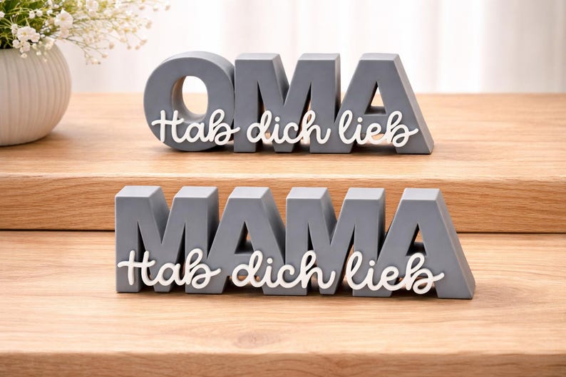 K&ouml;nnte beinhalten: Graue Holzbuchstabenbl&ouml;cke mit den W&ouml;rtern "OMA" und "MAMA" und dem wei&szlig;en Schriftzug "Hab dich lieb". Die Bl&ouml;cke stehen auf einer Holzoberfl&auml;che, im Hintergrund eine Vase mit wei&szlig;en Blumen.