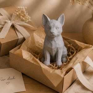 May include: A light gray, geometric, low-poly style dog figurine sits inside an open cardboard gift box lined with crinkled paper. A small card with the text "F&uuml;r dich" and a heart symbol is placed next to the box.