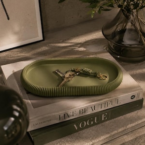 Puede incluir: Una bandeja ovalada verde oliva con bordes acanalados contiene llaves y un llavero. La bandeja se encuentra sobre una pila de libros, incluido uno titulado "LIVE BEAUTIFUL" y otro con la palabra "VOGUE". Un jarr&oacute;n con vegetaci&oacute;n est&aacute; en el fondo.