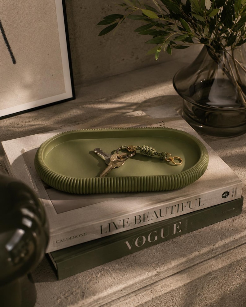 Puede incluir: Una bandeja ovalada verde oliva con bordes acanalados contiene llaves y un llavero. La bandeja se encuentra sobre una pila de libros, incluido uno titulado "LIVE BEAUTIFUL" y otro con la palabra "VOGUE". Un jarr&oacute;n con vegetaci&oacute;n est&aacute; en el fondo.