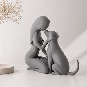 May include: A smooth, matte gray sculpture depicting a stylized figure kneeling and touching foreheads with a dog. The figure has long, flowing hair and a minimalist form, while the dog is shown in a seated position with its tail slightly raised.