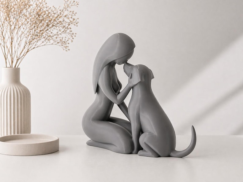 May include: A smooth, matte gray sculpture depicting a stylized figure kneeling and touching foreheads with a dog. The figure has long, flowing hair and a minimalist form, while the dog is shown in a seated position with its tail slightly raised.
