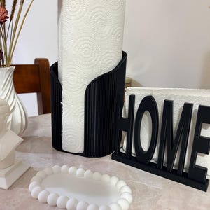 May include: A black vertical paper towel holder with a ribbed texture, a black napkin holder with the word HOME in capital letters, and a small white oval tray with a beaded edge. The paper towel roll has a white spiral pattern.