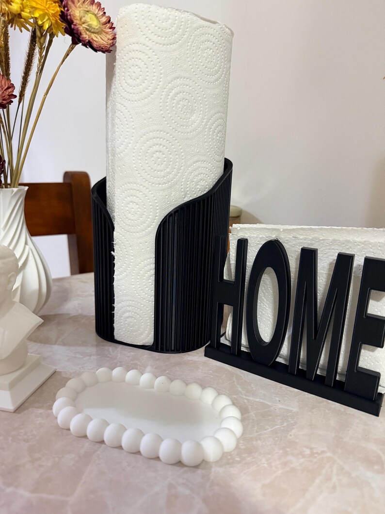 May include: A black vertical paper towel holder with a ribbed texture, a black napkin holder with the word HOME in capital letters, and a small white oval tray with a beaded edge. The paper towel roll has a white spiral pattern.