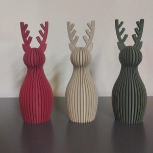 May include: Three standing reindeer figurines with a ribbed, vertical slat construction. The left figure is deep red, the center figure is off-white, and the right figure is dark olive green. Each has a rounded body and branched antlers.